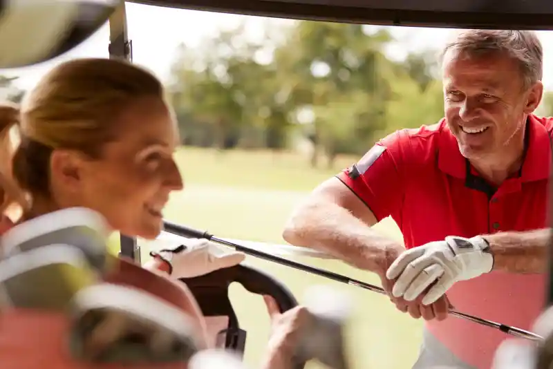 A man in a red polo shirt smiles while leaning on a golf cart, engaging with a woman who is seated in the cart, wearing golf gloves. Green grass and trees are visible in the background.