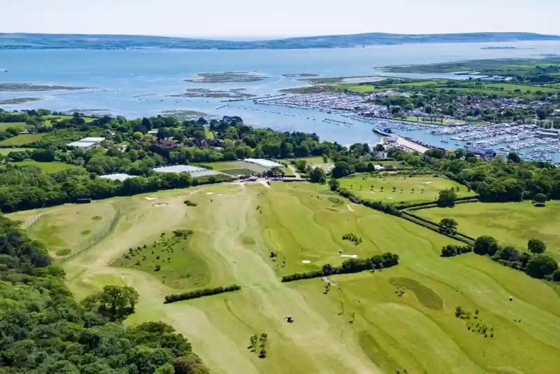 Aerial view of a lush green landscape featuring a golf course, with a marina and boats in the background. The coastline and calm blue waters are visible, alongside patches of trees and fields.