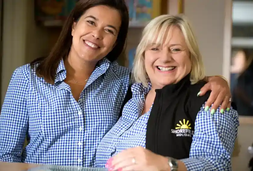 Two smiling women stand together, both wearing checked blue shirts. One woman has long dark hair, while the other has short blonde hair and wears a black vest with the logo of Shorefield Holidays; They appear friendly and welcoming.