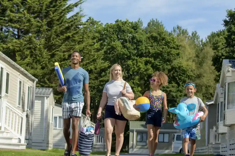 A family walks along a pathway lined with holiday homes. They carry beach toys, towels, and other supplies, with greenery in the background and a clear blue sky above.