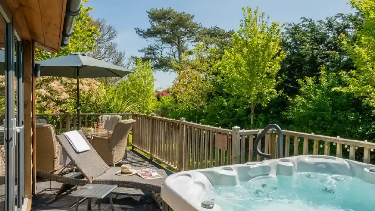 A cosy outdoor deck featuring a hot tub, sun loungers, and an umbrella. Lush greenery surrounds the area, creating a serene atmosphere.