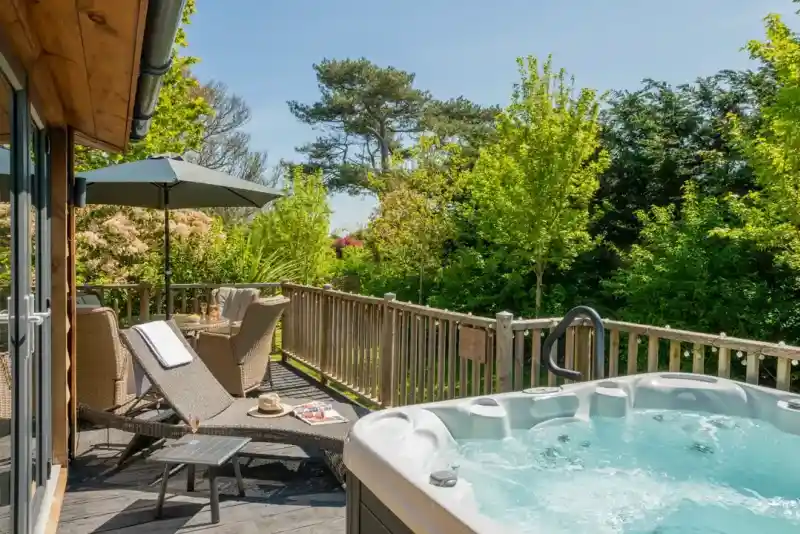 A cosy outdoor deck featuring a hot tub, sun loungers, and an umbrella. Lush greenery surrounds the area, creating a serene atmosphere.