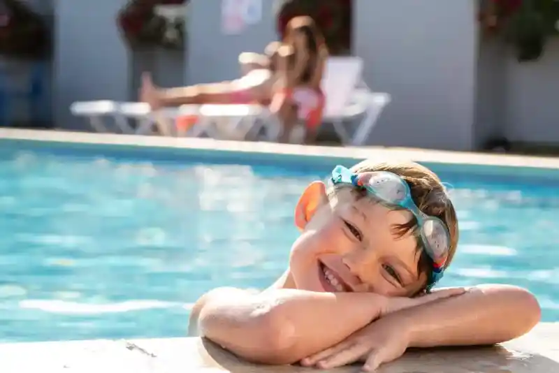 A young boy with swim goggles smiles while leaning over the edge of a swimming pool. In the background, a woman relaxes on a lounge chair.