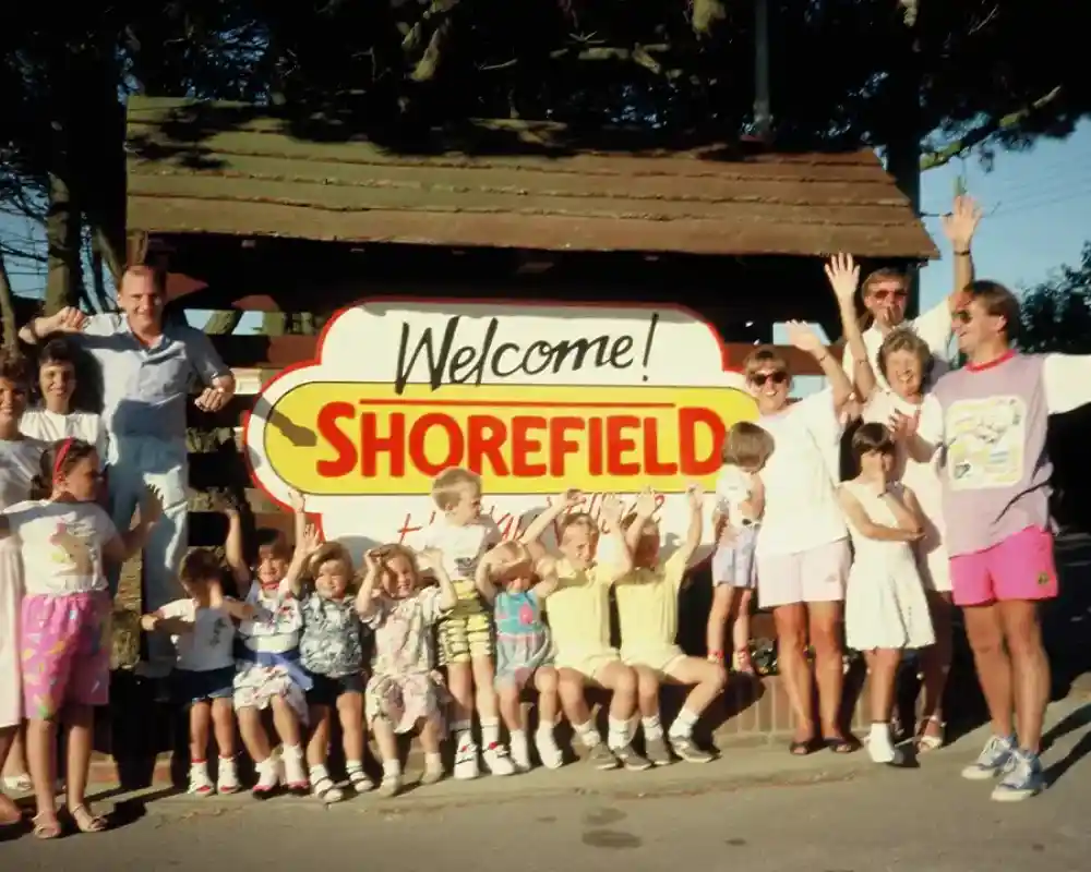 A large group of people, including children and adults, gather around a colourful Welcome to Shorefield sign. Many are smiling and waving, exuding a festive atmosphere. The background features trees and a clear sky.