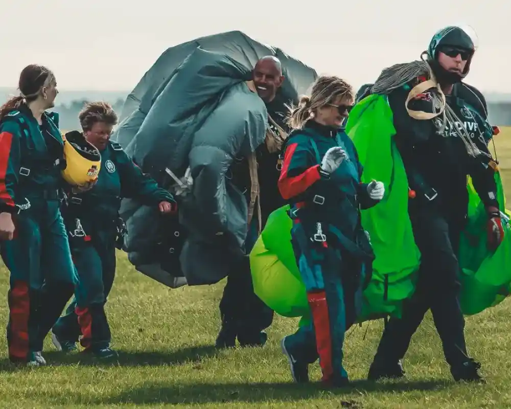A group of skydivers, dressed in colourful jumpsuits and harnesses, walk across a grassy field. They are carrying parachutes, and some appear to be smiling and chatting with each other. The background features a clear sky and distant hills.