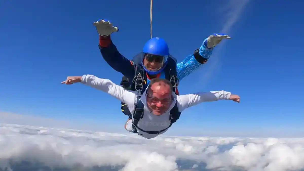 Two skydivers are in freefall, with one person in front and the other behind, both wearing parachute gear and helmets. They are soaring above a cloudy landscape, with a bright blue sky in the background.
