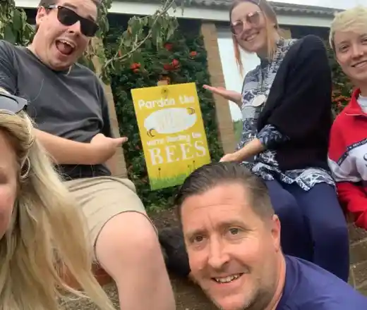 A group of five people pose together outdoors, smiling and pointing at a sign that says Pardon the weeds, we're feeding the bees. They are seated on a low wall with greenery in the background.
