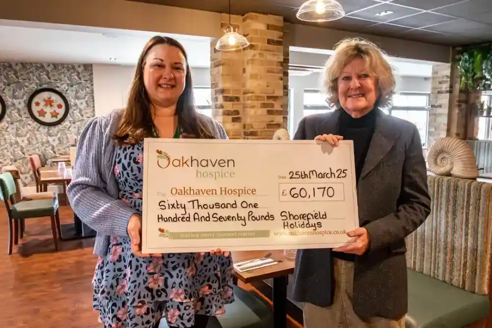 Two women stand in a café, holding a large check for £60,170 made out to Oakhaven Hospice. The check is dated March 25. The background features a rustic stone wall and comfortable seating.