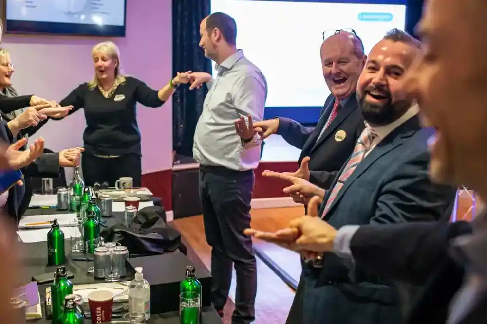 A group of professionals stands in a conference setting, smiling and interacting enthusiastically. Some individuals are gesturing with their hands, while others appear to be engaged in a light-hearted activity. Tables in the foreground are set with green bottles and glasses.
