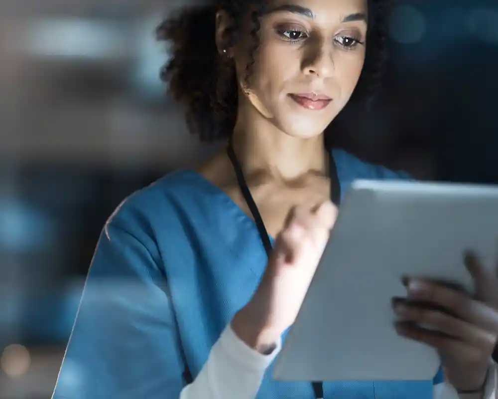 A healthcare professional in scrubs uses a tablet while standing in a hospital setting. She appears focused and engaged. A hospital ID badge is visible around her neck.
