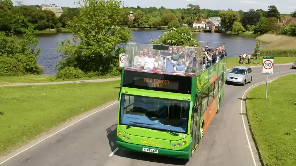 A green and orange double-decker bus with an open-top is driving along a road next to a lake. Passengers are enjoying the view as trees and houses are visible in the background under a clear blue sky.