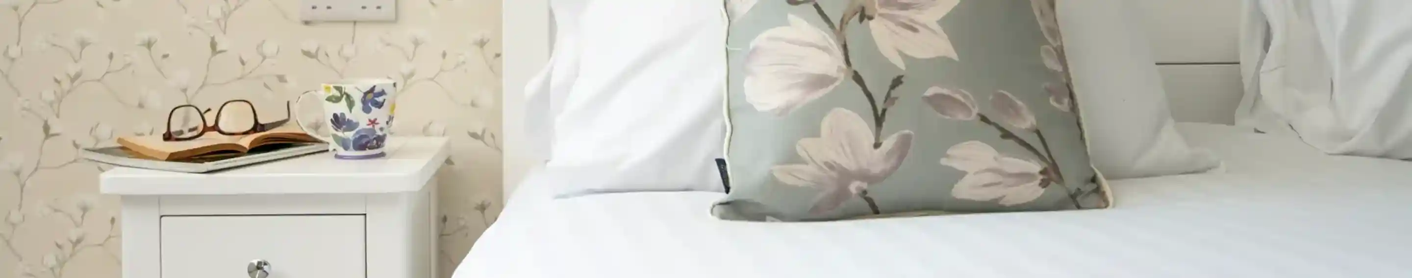 A neatly made bed with white linens and a decorative floral pillow. A small nightstand holds a pair of glasses, an open book, and a floral mug. The background features light-coloured, patterned wallpaper.