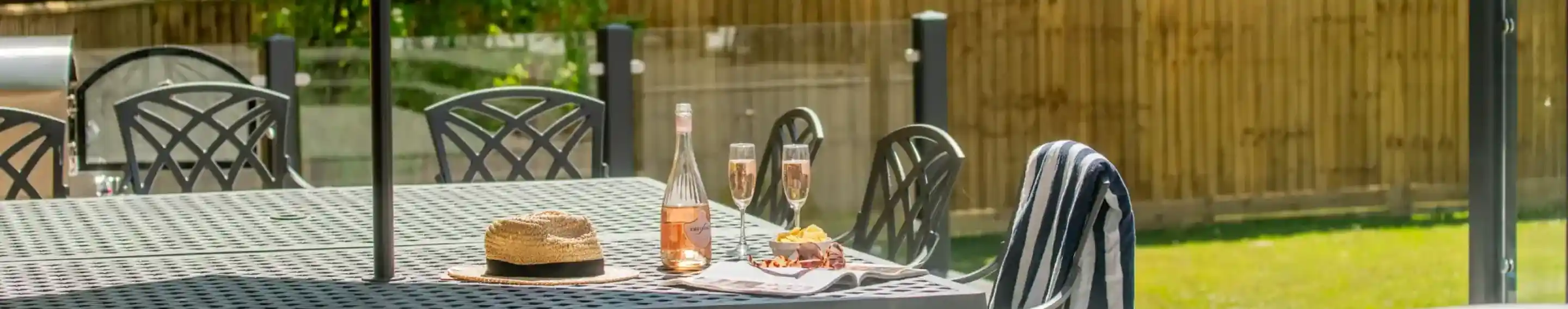 A relaxed outdoor setting featuring a patio table with a bottle of rosé, two champagne flutes, and a plate of snacks. There is a straw hat on the table, and a hot tub is visible in the foreground, with greenery and a wooden fence in the background.