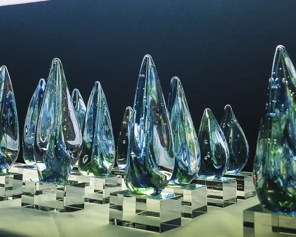 Clear glass awards in the shape of teardrops are displayed on a table. A banner in the background reads "Welcome to Excellence." The setting is elegant, with soft lighting that highlights the awards.