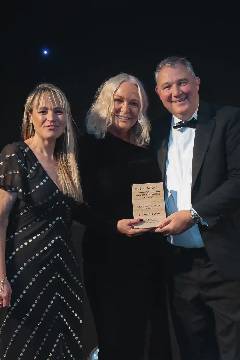 Three individuals pose together at an award ceremony. One woman, wearing a black dress with white polka dots, stands on the left, while a second woman in a black outfit holds a plaque in the centre. A man in a tuxedo stands on the right, smiling widely. The backdrop is decorated with subtle lighting.