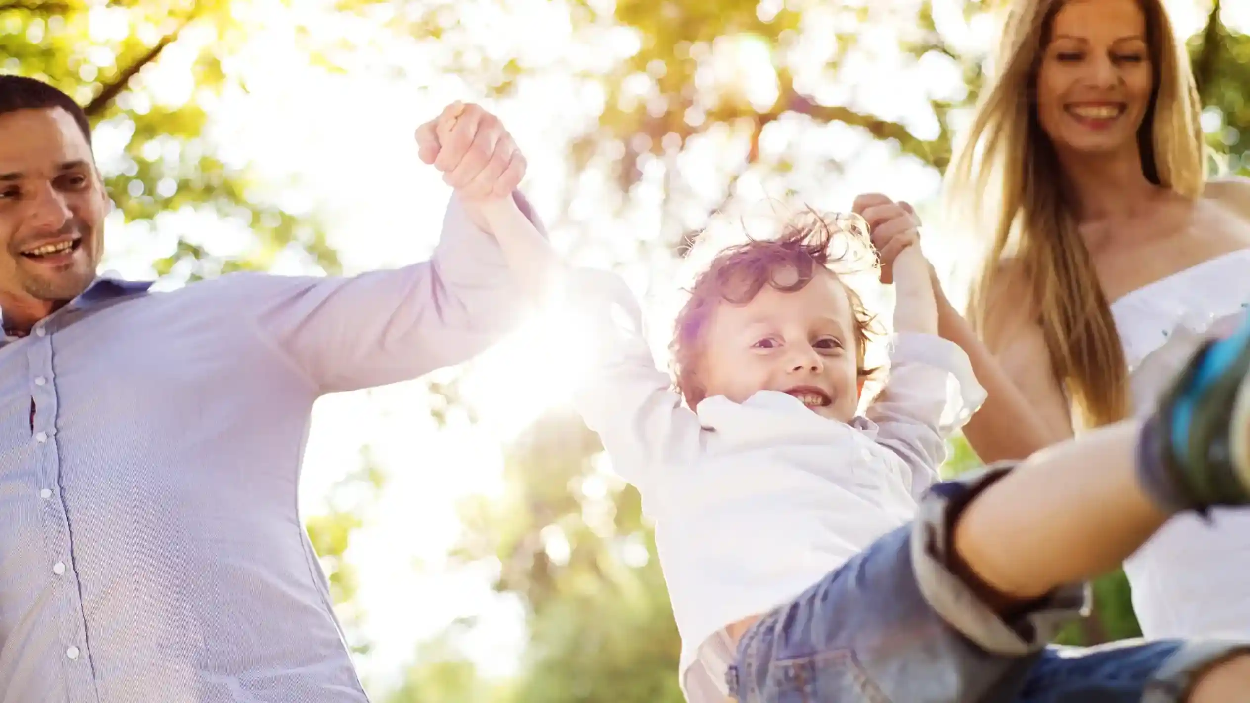 A joyful child swings between two smiling adults, holding hands and enjoying a sunny day in a park with green trees in the background.