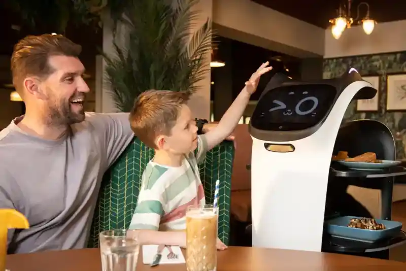 A smiling man sits at a table in a restaurant with a young boy, who is playfully interacting with a robotic server. The robot has a screen displaying a friendly face and is presenting food on its shelves. The setting features decorative plants and a cosy atmosphere.