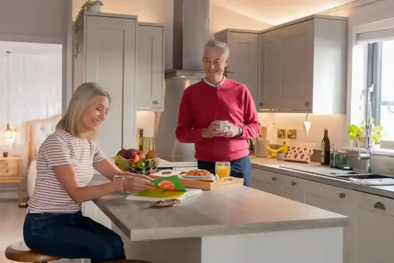 A woman sitting at a kitchen island smiles while reading a card, while a man stands nearby holding a mug and looking at her with a smile. The kitchen has modern cabinetry, and a fruit bowl and snacks are on the island.