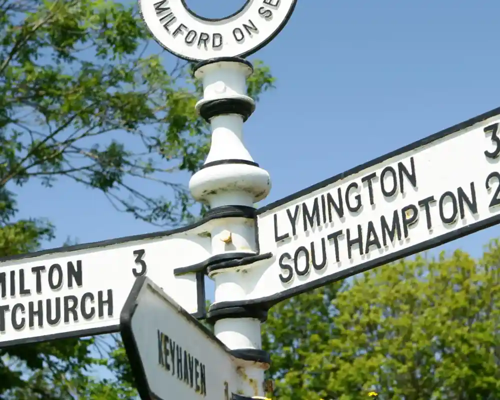 Signpost indicating directions and distances to Milton, Lymington, Southampton, and Keyhaven, surrounded by colourful flowers under a blue sky.