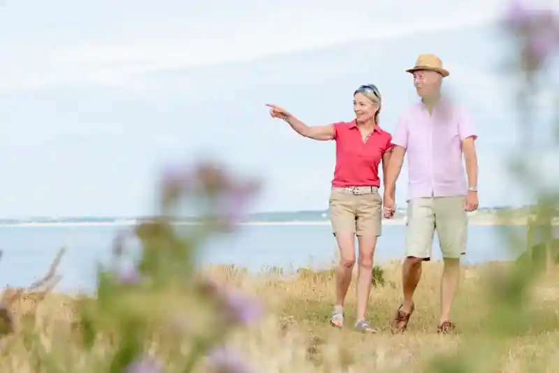 A couple strolls hand in hand along a scenic coastal path, with the woman pointing towards the horizon. They are dressed in casual summer attire, and the landscape features grasses and blooming flowers.