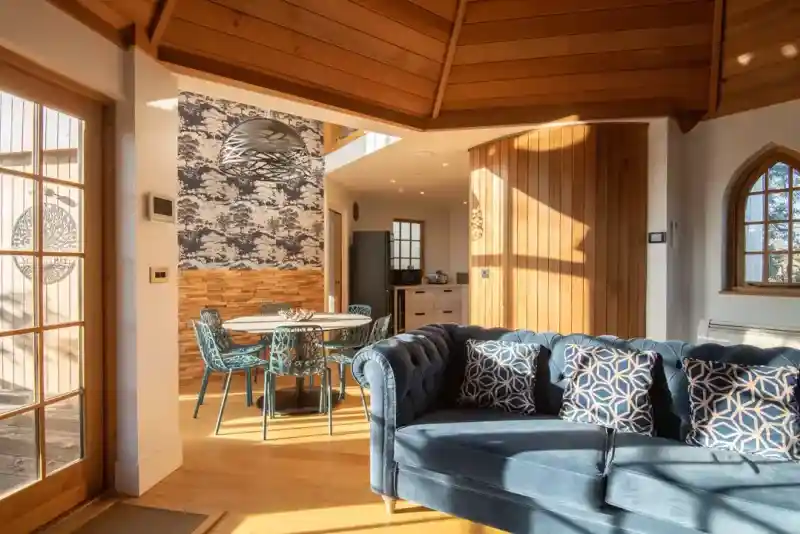 A cozy interior space featuring a blue tufted sofa with decorative cushions, a round dining table with patterned chairs, and a well-lit area with wooden beams and unique wall decor. Natural light streams through large windows.