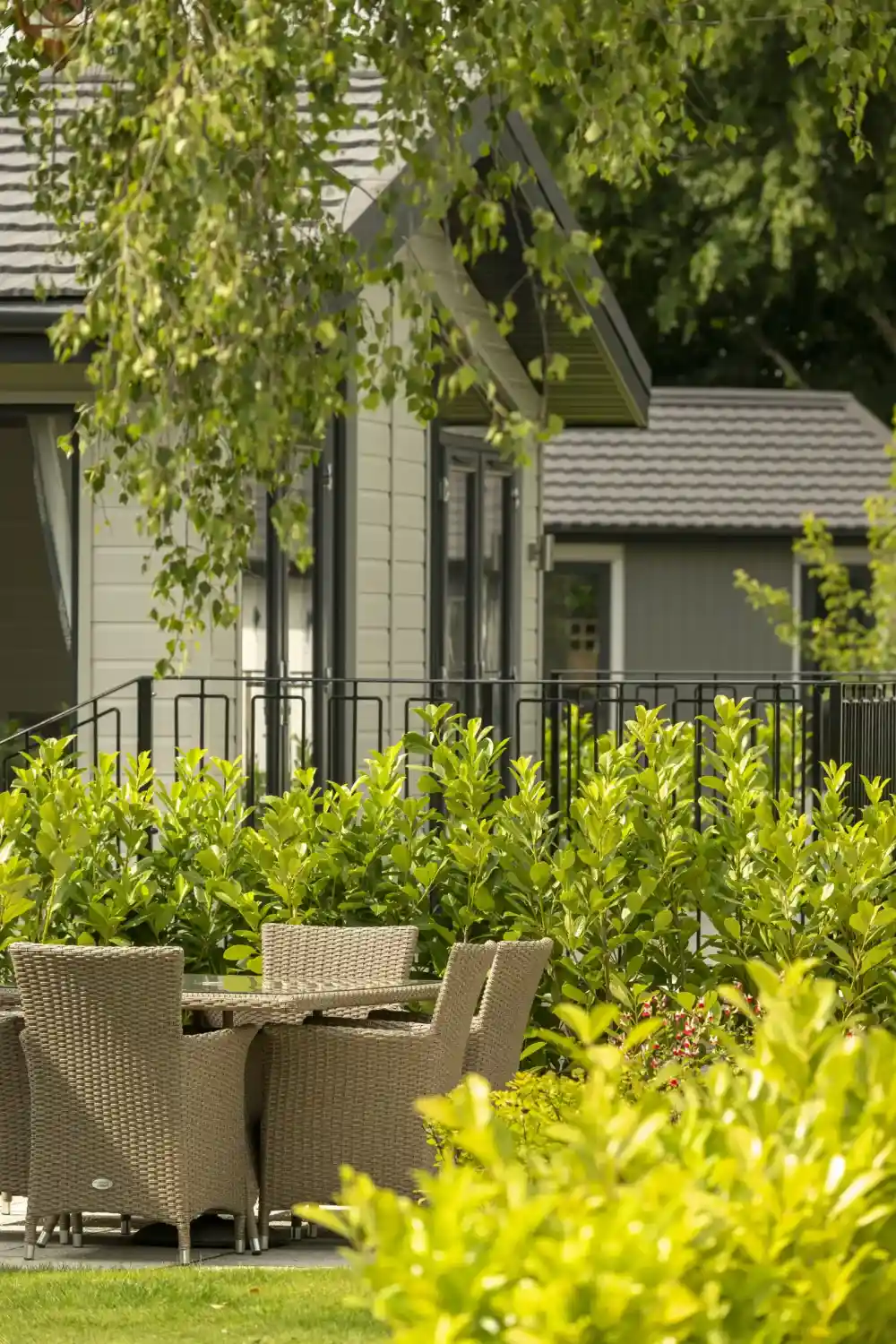 A cosy outdoor seating area with a round table and four wicker chairs surrounded by lush green bushes. In the background, there are two modern cabin-like buildings with dark roofs and large windows framed by trees.