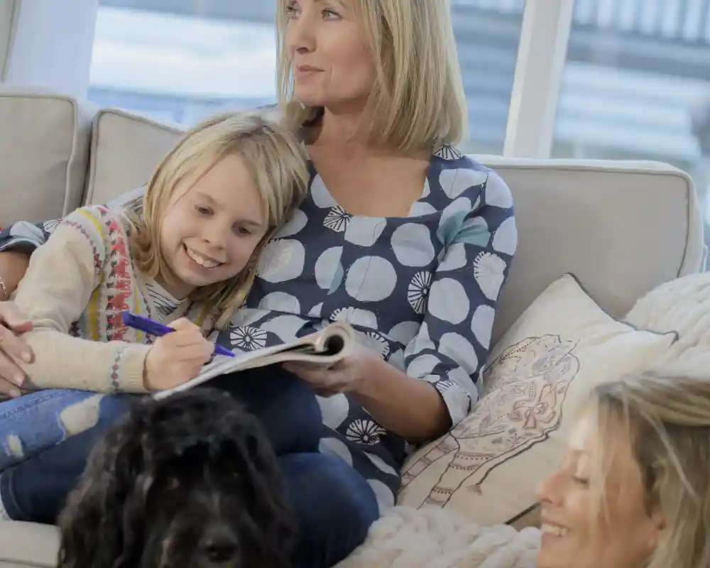 A woman sits on a couch, smiling while a young girl leans against her, drawing in a notebook. Another woman sitting nearby looks on with a smile. A black dog lies on the floor, adding warmth to the cosy, bright living room scene.