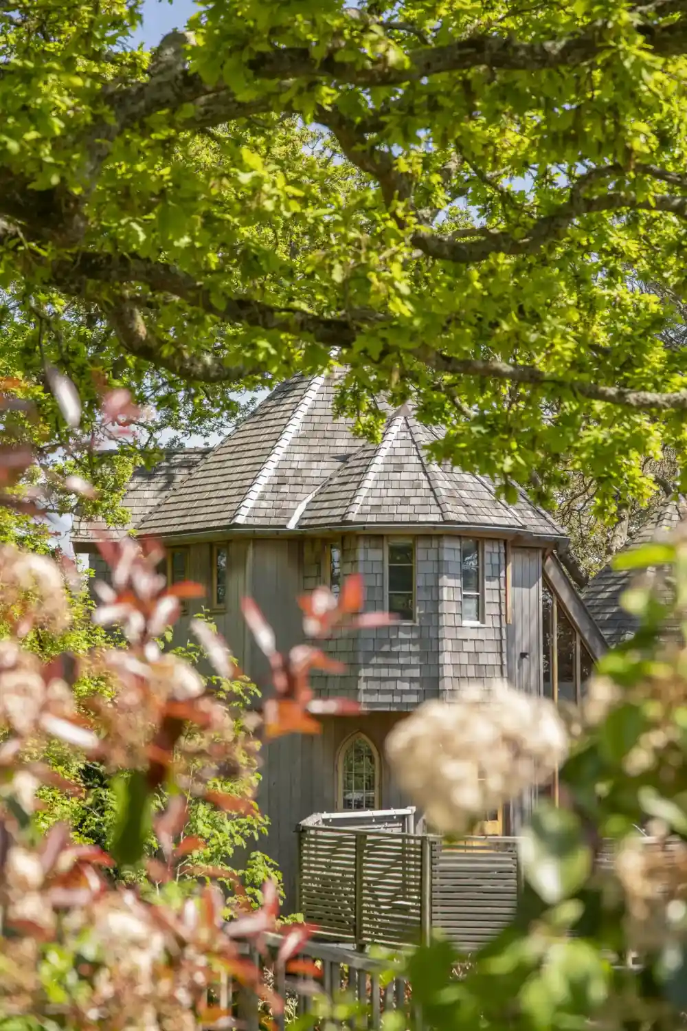 A charming wooden house with a unique conical roof, surrounded by lush green trees and colourful flowering plants. Sunlight filters through the leaves, creating a warm and inviting atmosphere.