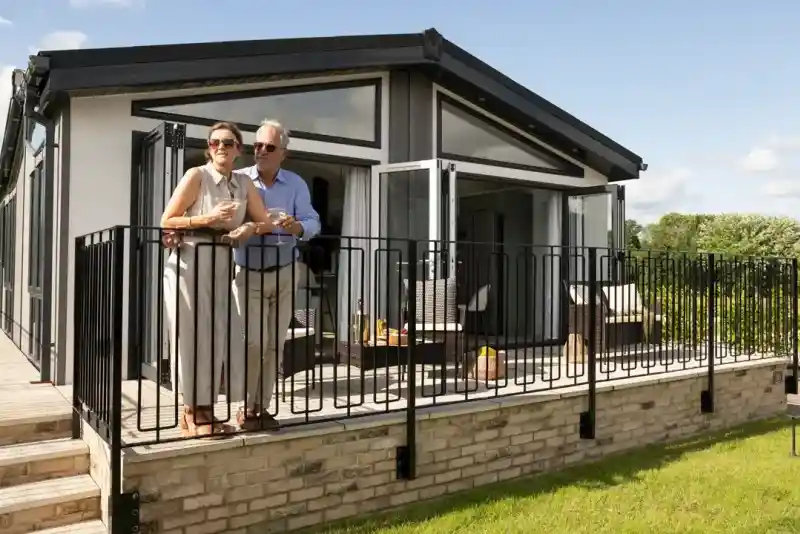 A couple stands on a balcony of a modern house, enjoying drinks and looking out at the landscape. The house features large glass windows and a sleek design, with greenery in the background.