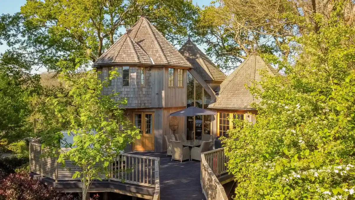 A charming wooden house with a unique multi-pitched roof, surrounded by lush green trees and foliage. A deck with outdoor furniture is visible in front, invitingly situated under a shaded area.