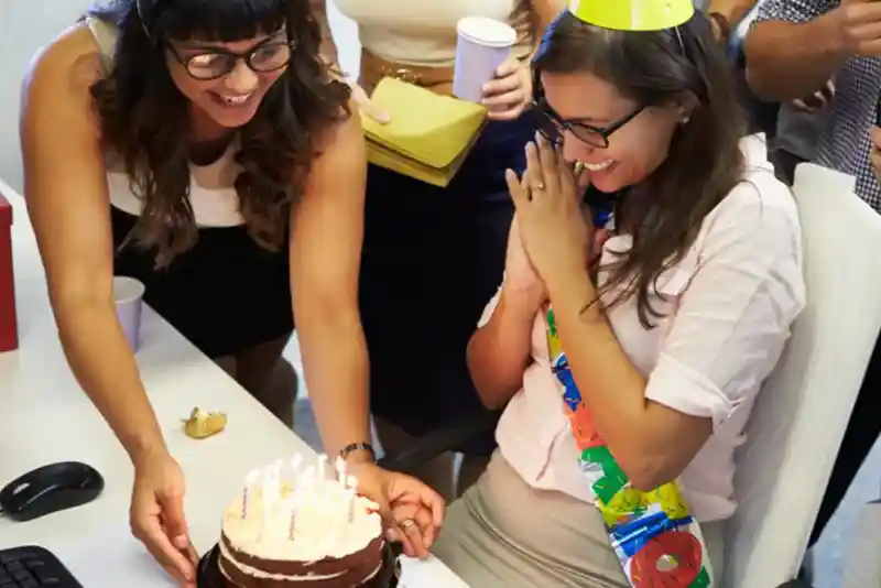 Two women celebrate a birthday at an office. One woman lights candles on a chocolate cake while the other, wearing glasses and a party hat, looks excitedly at the cake with hands clasped together. Friends gather around, holding cups and smiling.