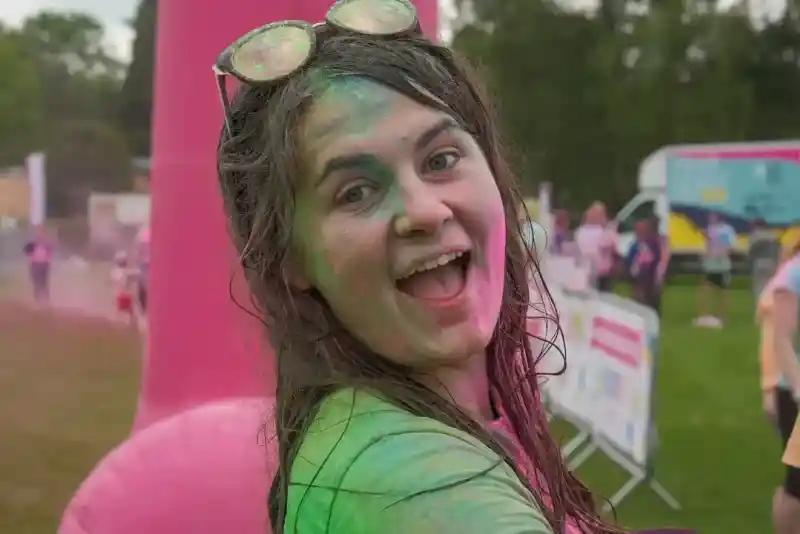 A young woman with wet hair and colourful powder on her face smiles widely, standing in a festive outdoor setting. She wears sunglasses on her head and is surrounded by blurred figures in the background.