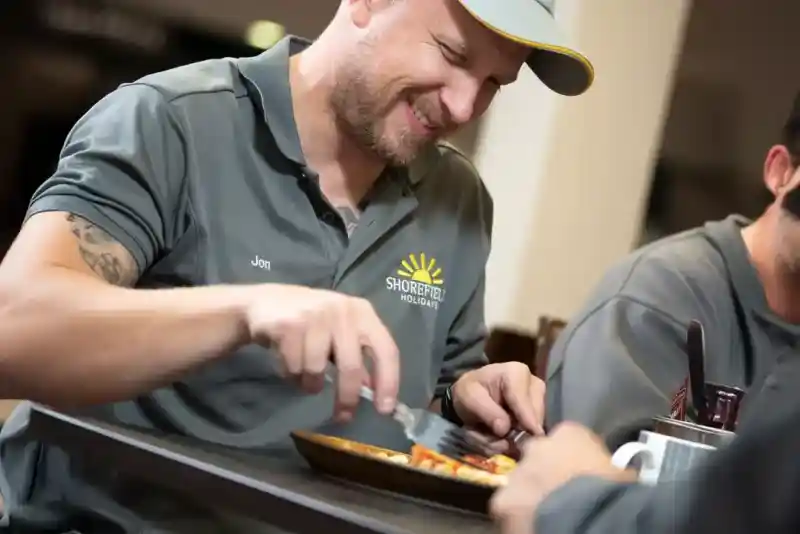 A man in a grey shirt with a logo smiles while enjoying a meal at a table. He leisurely cuts into his food, with other diners visible in the background.