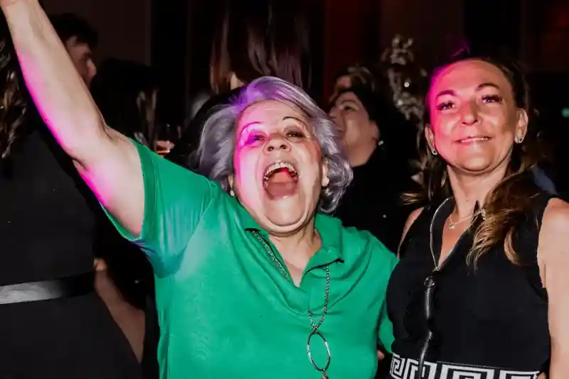 Two women enjoying a lively moment at a party. One woman, wearing a green shirt, is energetically raising her arms and laughing, while the other, dressed in black, smiles beside her. The background features colourful lighting and other party attendees.