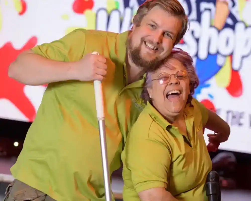 Two smiling individuals wearing matching green shirts pose playfully together. One holds a cleaning tool while the other leans in with a joyful expression. A colourful, cheerful backdrop featuring the woofs Workshop is visible behind them.