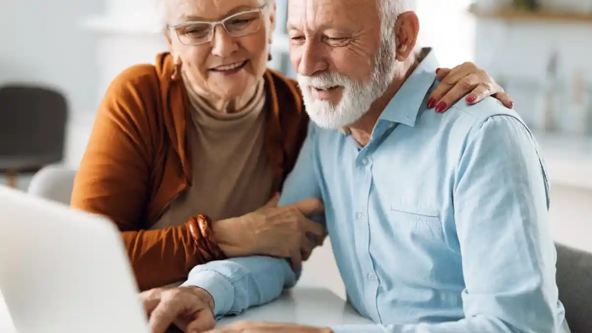 An elderly couple smiles together while sitting at a table, looking at a laptop. The woman has glasses and light grey hair, wearing a brown cardigan, while the man has a full beard and light grey hair, dressed in a blue shirt. They appear to be enjoying each other's company.