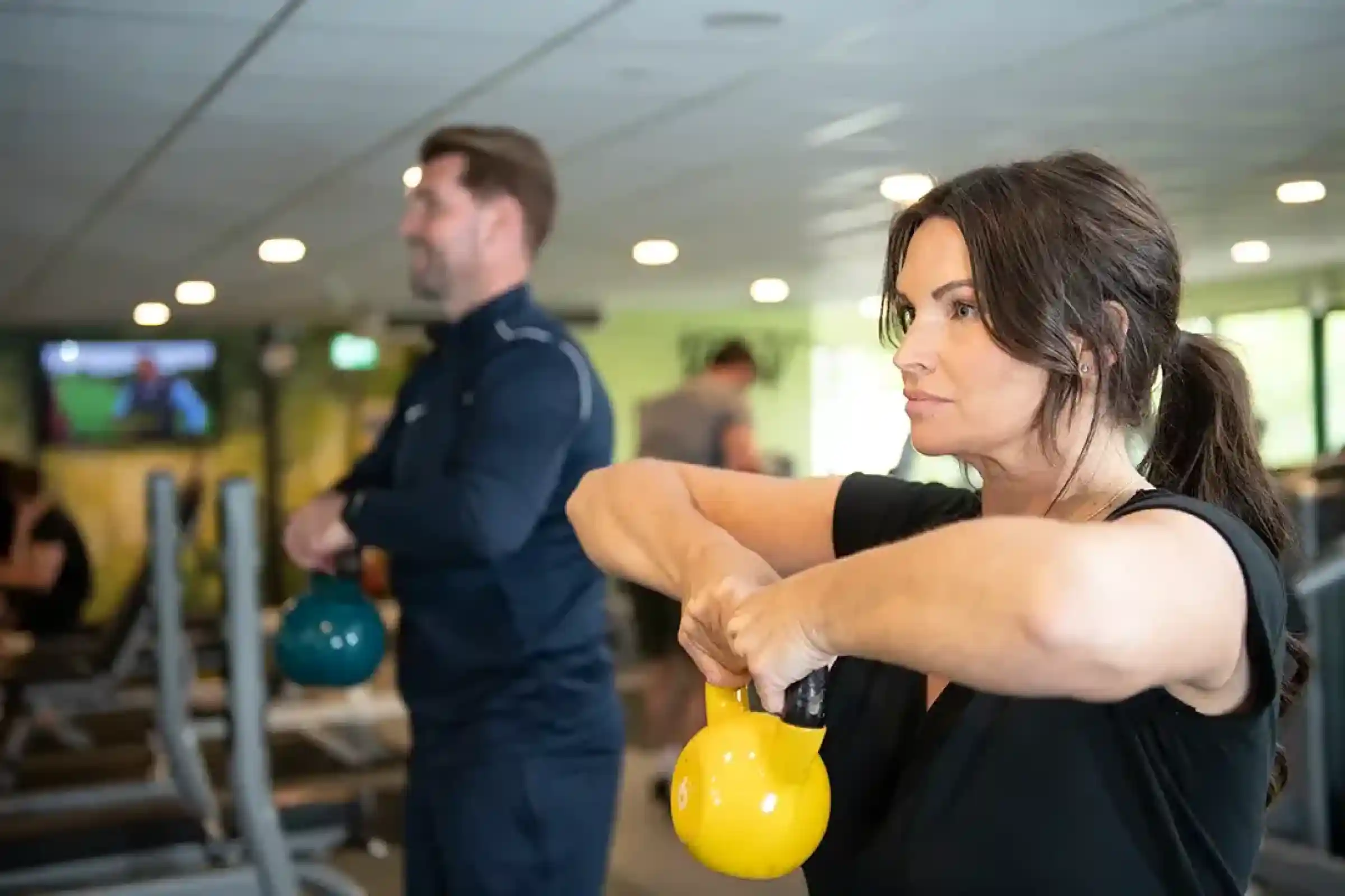 A woman and a man are exercising in a gym, both holding kettlebells. The woman is focused and performing a kettlebell squat, while the man is seen in the background, also engaged in a workout. The gym has various fitness equipment and a TV screen displaying content.