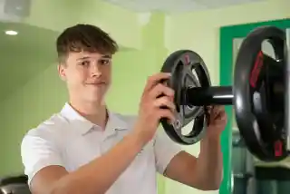A young man in a white polo shirt adjusts weights on a barbell in a gym. He is smiling and appears focused on his task. The gym background features green walls and workout equipment.