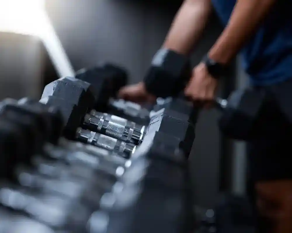A person lifting a dumbbell from a rack in a gym, showcasing various weights arranged neatly. The focus is on the individual and the dumbbell, with a blurred background highlighting the gym environment.