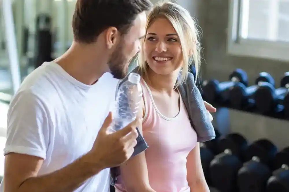 A smiling man and woman are sitting close together in a gym, engaged in a lighthearted conversation. The man is holding a water bottle, while the woman has a towel draped over her shoulder. In the background, gym equipment is visible.