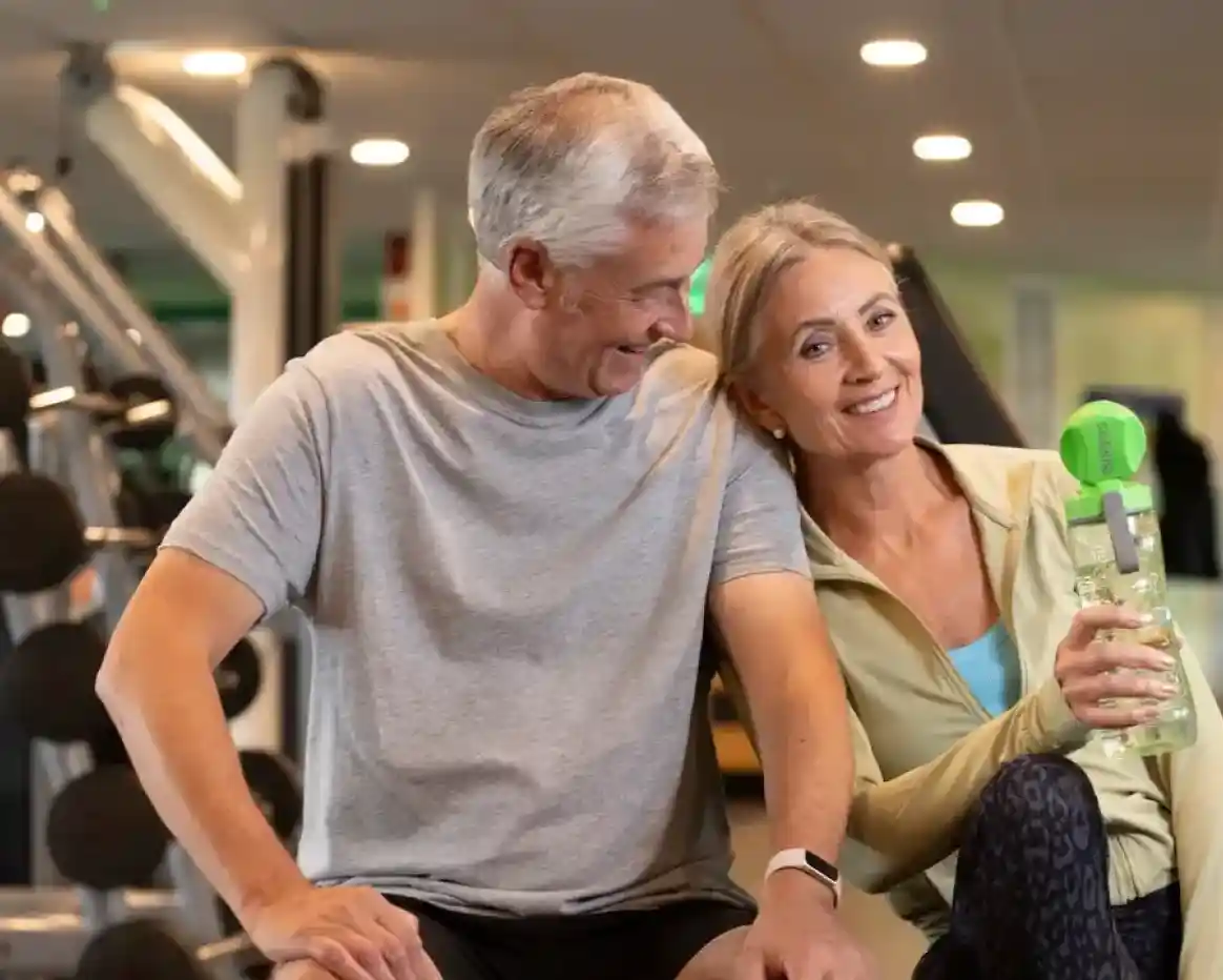A smiling older couple sits together in a gym. The man, in a gray shirt, looks at the woman, who holds a green-top water bottle. They appear relaxed and happy.