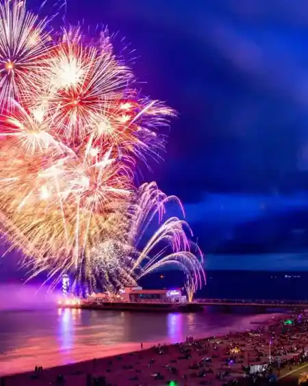 Colorful fireworks explode in the night sky above a beach, illuminating the dark clouds and reflecting on the water. People gather on the shoreline, enjoying the vibrant display near a pier.