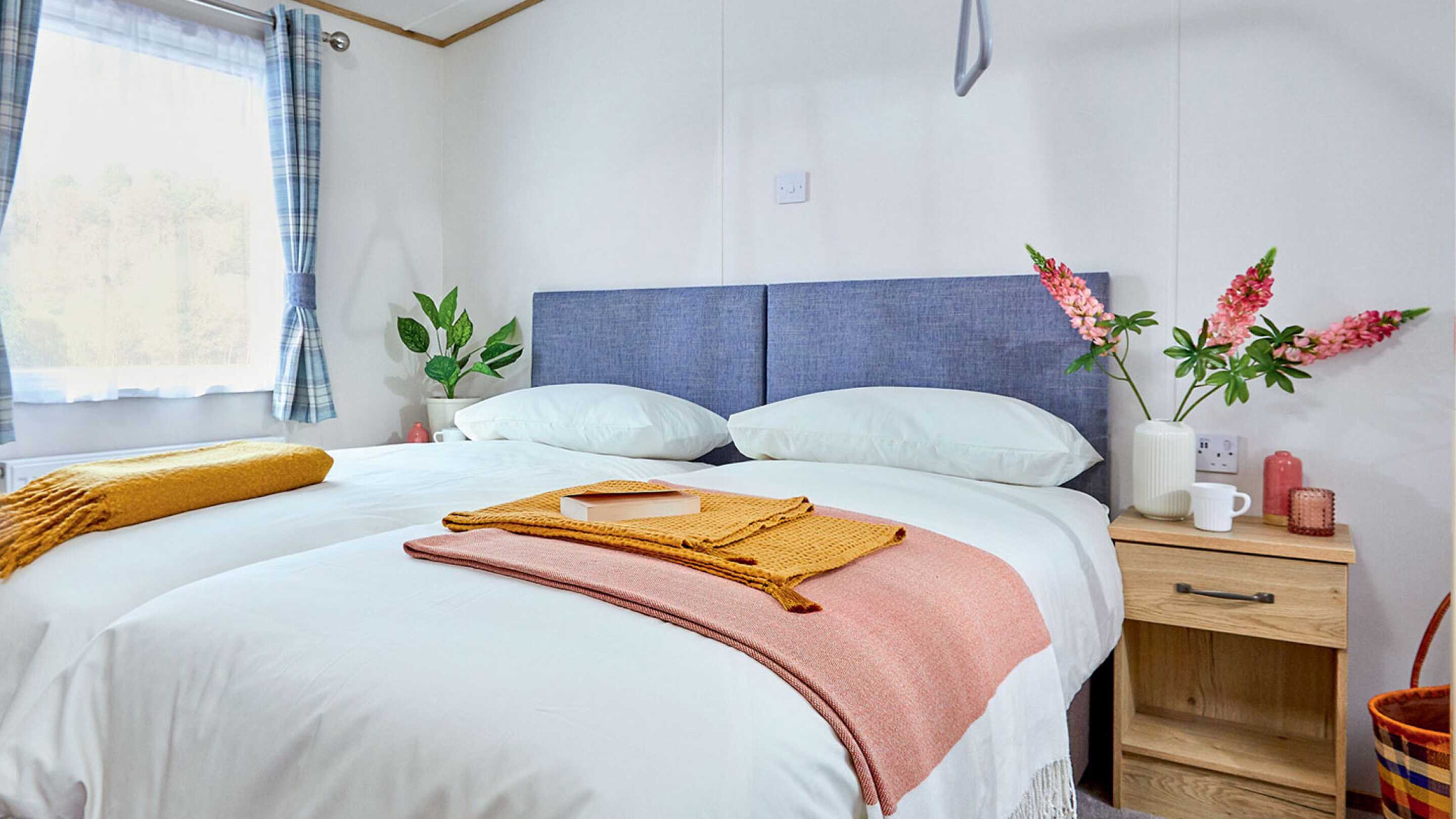 A cozy bedroom featuring two neatly made beds with blue headboards. A colorful throw blanket is draped over one bed, and a bedside table holds a small decorative item. Fresh flowers in a vase add a touch of color, and sunlight filters through the window, illuminating the space.