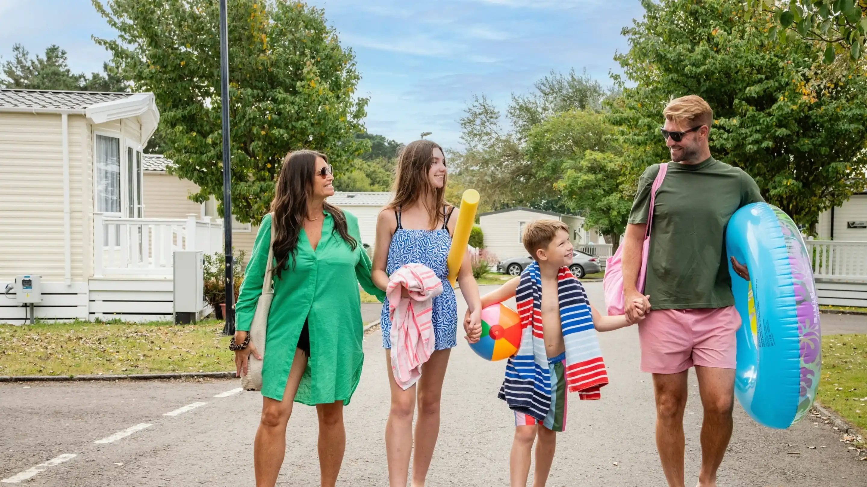 Four people walk together along a park pathway, smiling and holding beach gear. Two women wear colorful summer attire, while a boy in the middle holds a water toy, and a man carries a large inflatable float. Lush trees and holiday homes line the background.