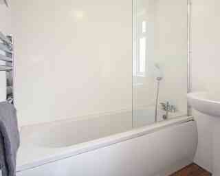 A modern bathroom featuring a white bathtub with a glass shower screen, a small sink, and a towel hanging on a rack. The walls are painted white, and the space is well-lit with natural light.