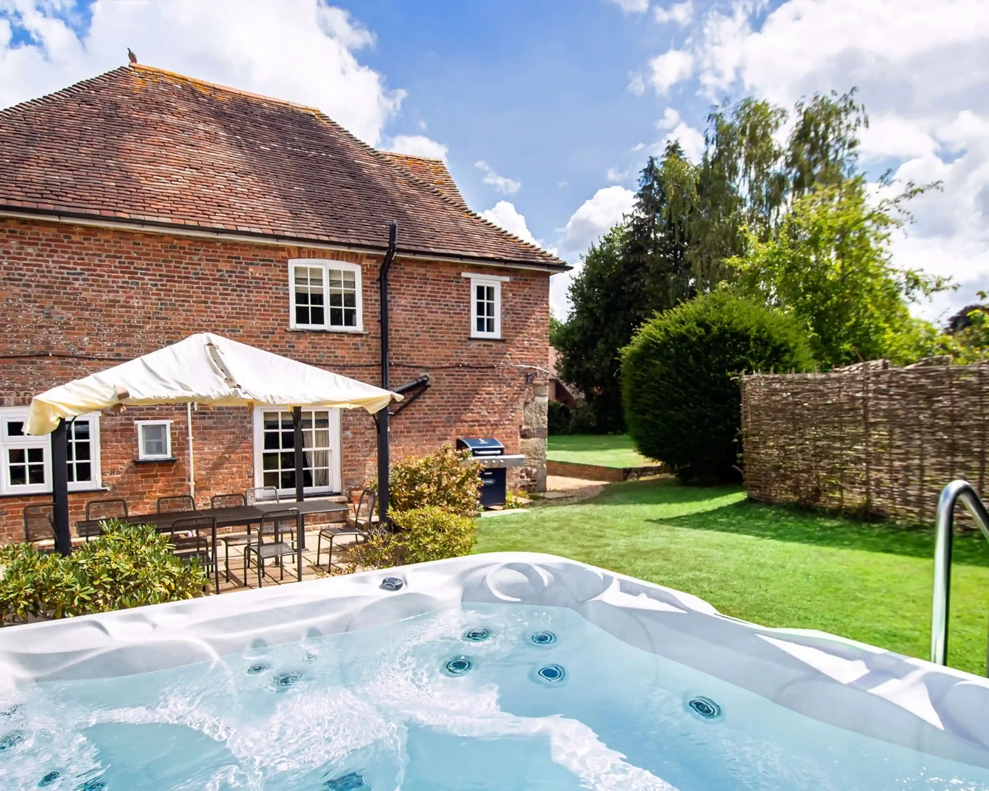 A hot tub with bubbling water is in the foreground, surrounded by a grassy area. In the background, a brick house with a sloped roof and white windows is visible, along with a shaded seating area. Lush greenery and a stone wall enhance the peaceful outdoor setting under a partly cloudy sky.