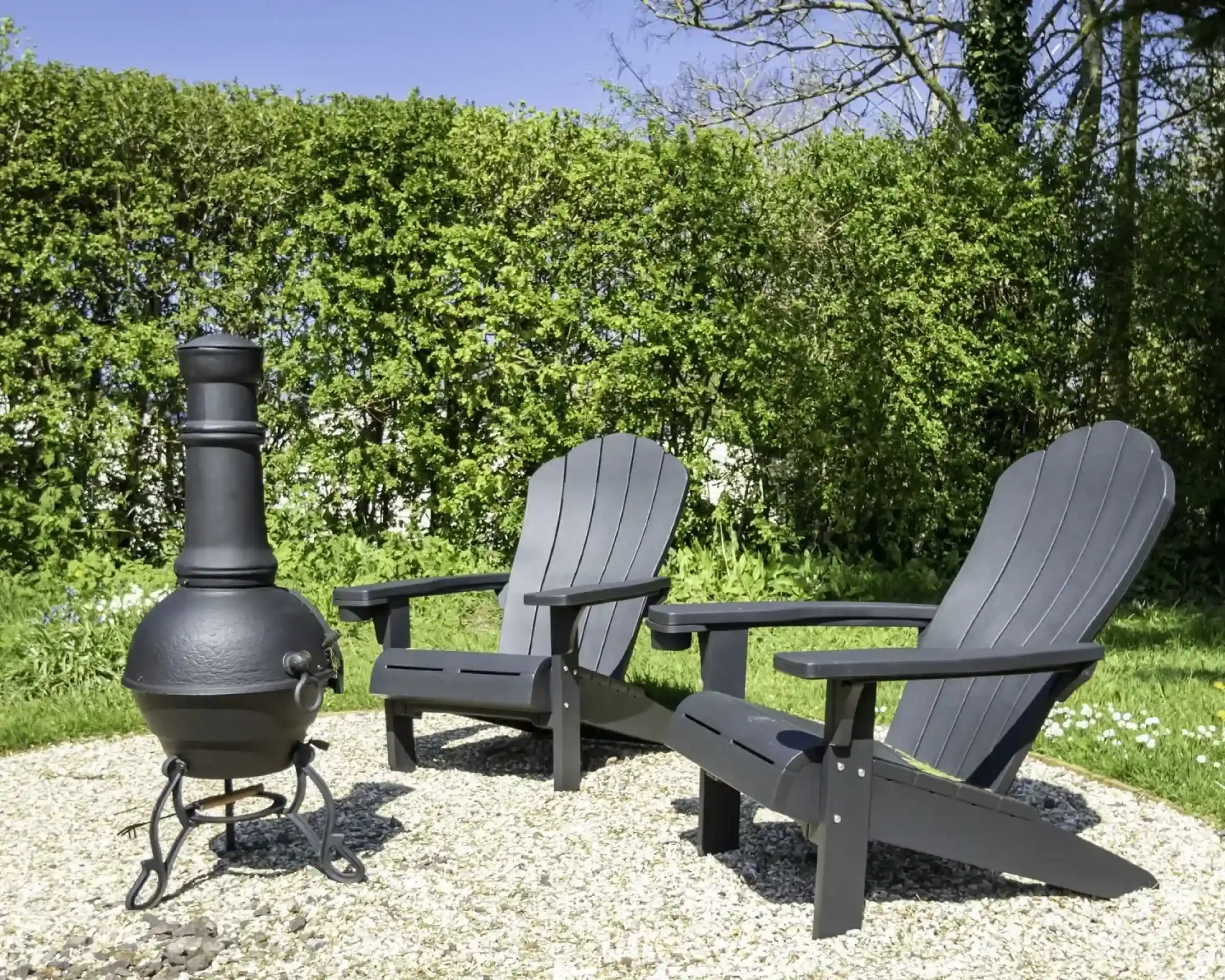Two black Adirondack chairs are positioned on a gravel area, beside a traditional cast iron chiminea. Lush green hedges and a blue sky serve as the backdrop. White flowers are scattered on the ground.