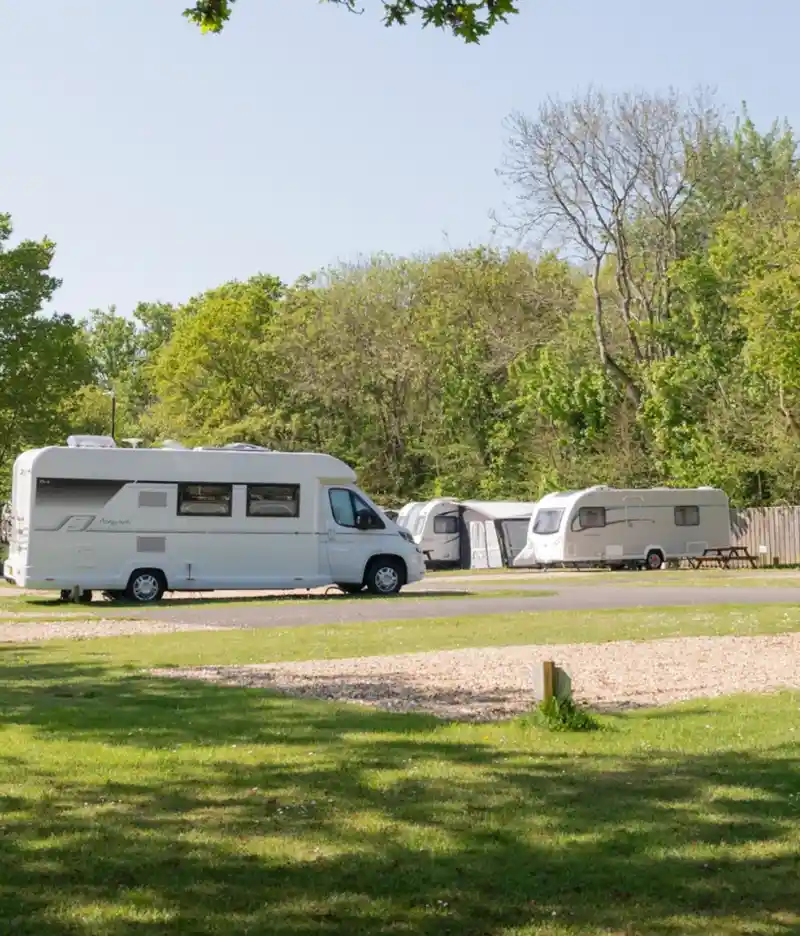 Aerial view of a campsite with grassy areas, trees, and several parked caravans. In the background, the coastline is visible along with a distant town and fields. Clear blue sky enhances the scenic landscape.