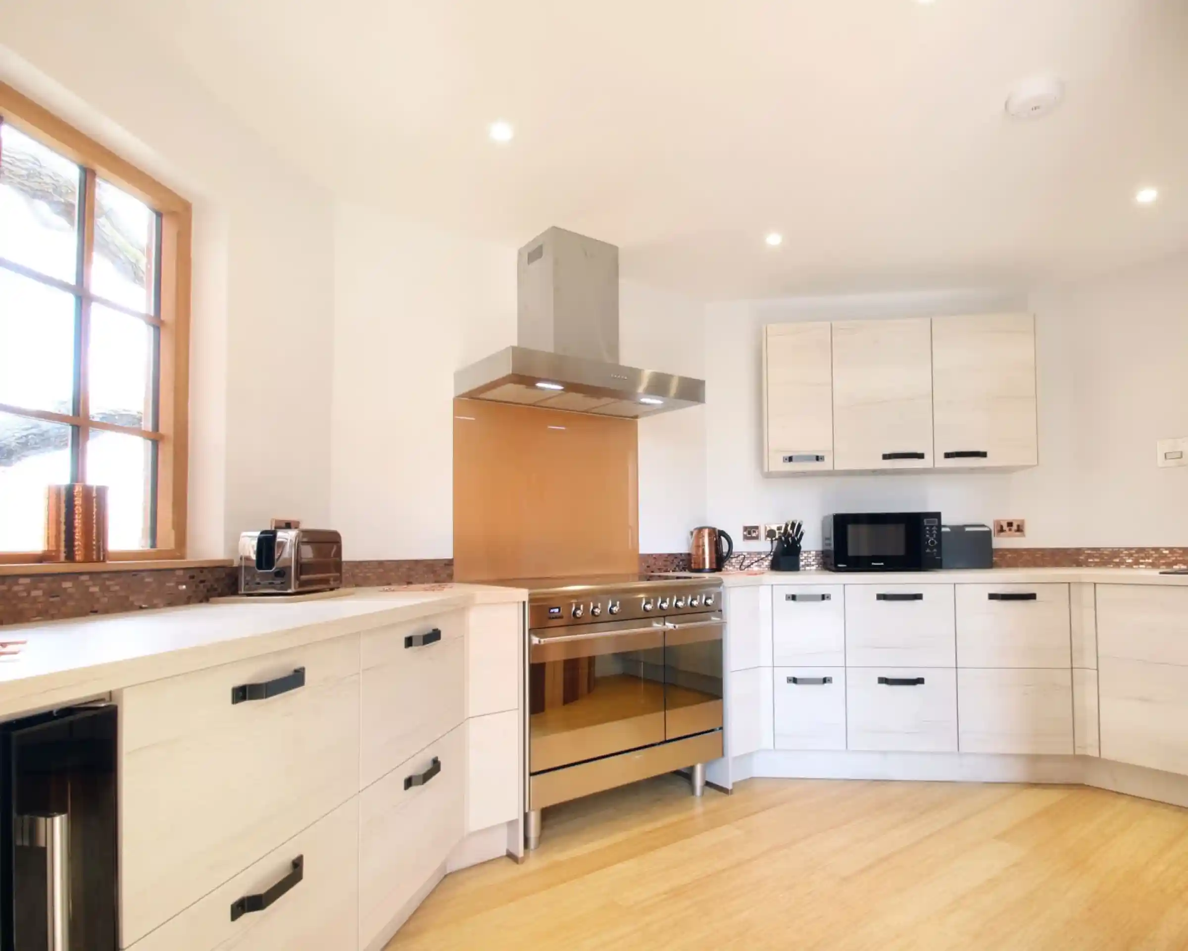 A modern kitchen featuring light-colored cabinets, a sleek golden stovetop, and stainless steel appliances. Large windows allow natural light to brighten the space, complemented by warm wooden flooring.