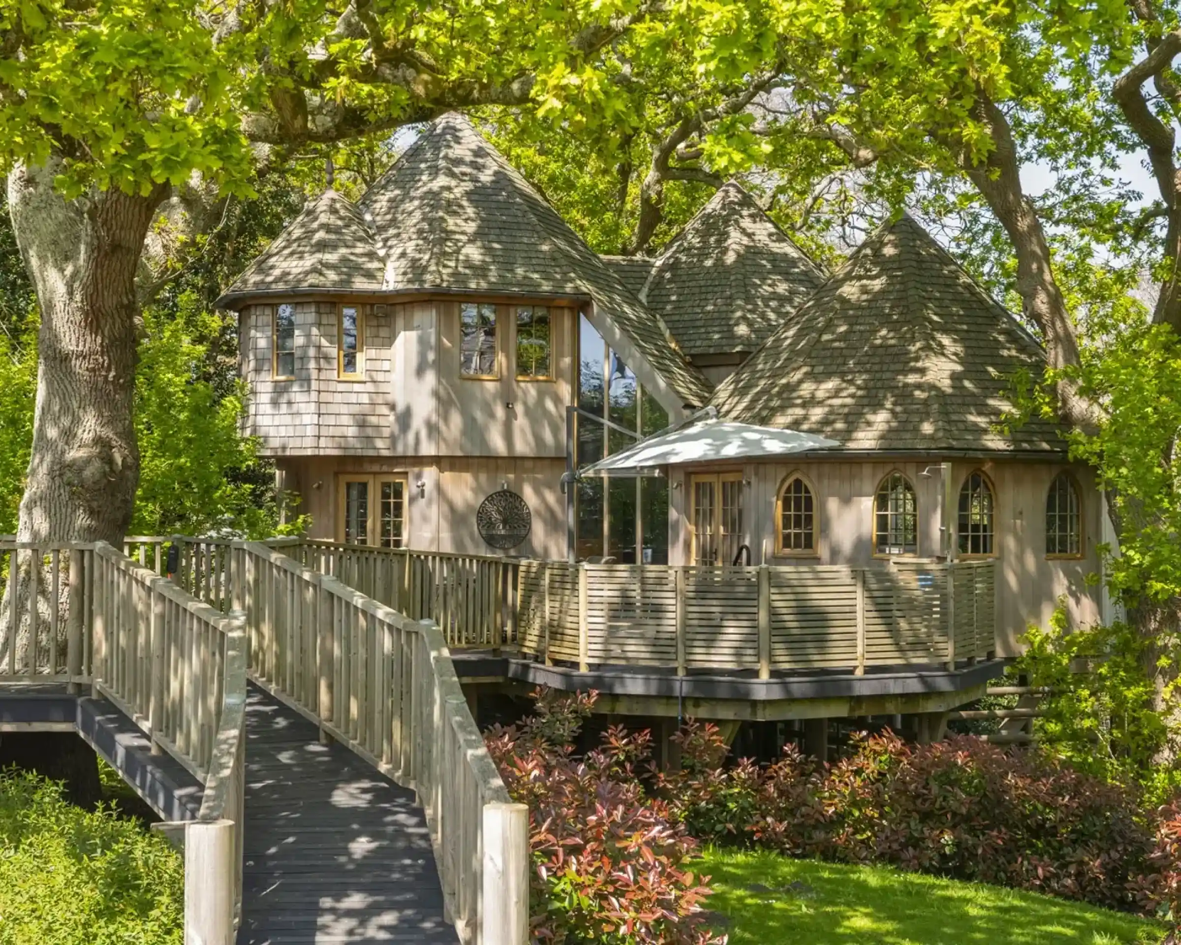 A charming wooden house with a unique, rounded architecture sits among lush greenery. It features a wraparound deck, large windows, and a sloping roof. A wooden walkway leads to the entrance, surrounded by vibrant plants and trees.